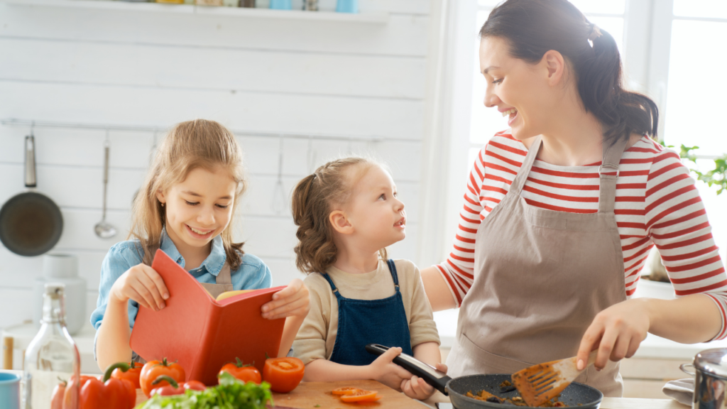 A mom cooking with her two daughters, one reading what's meant to be a cookbook, the other exchanging a look with her mom
